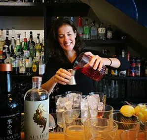 Vrouw maakt cocktails achter een bar met flessen en glazen op de voorgrond.