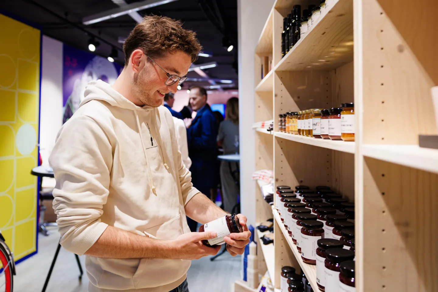 Bezoeker bekijkt een potje jam in de Makerswinkel van ROC Midden Nederland, met houten schappen vol producten en andere bezoekers op de achtergrond.