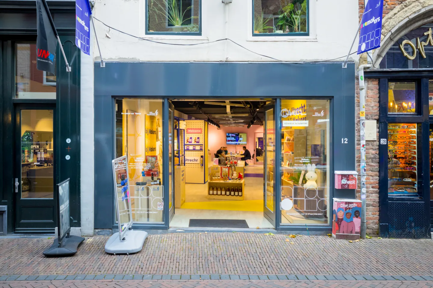 Ingang van de winkel Makers aan een gezellige winkelstraat in Utrecht. De glazen deuren staan open en binnen zijn houten displays met lokale producten te zien. Aan de gevel hangen blauwe vlaggen met het Makers-logo, en voor de deur staan reclameborden op de stoep.