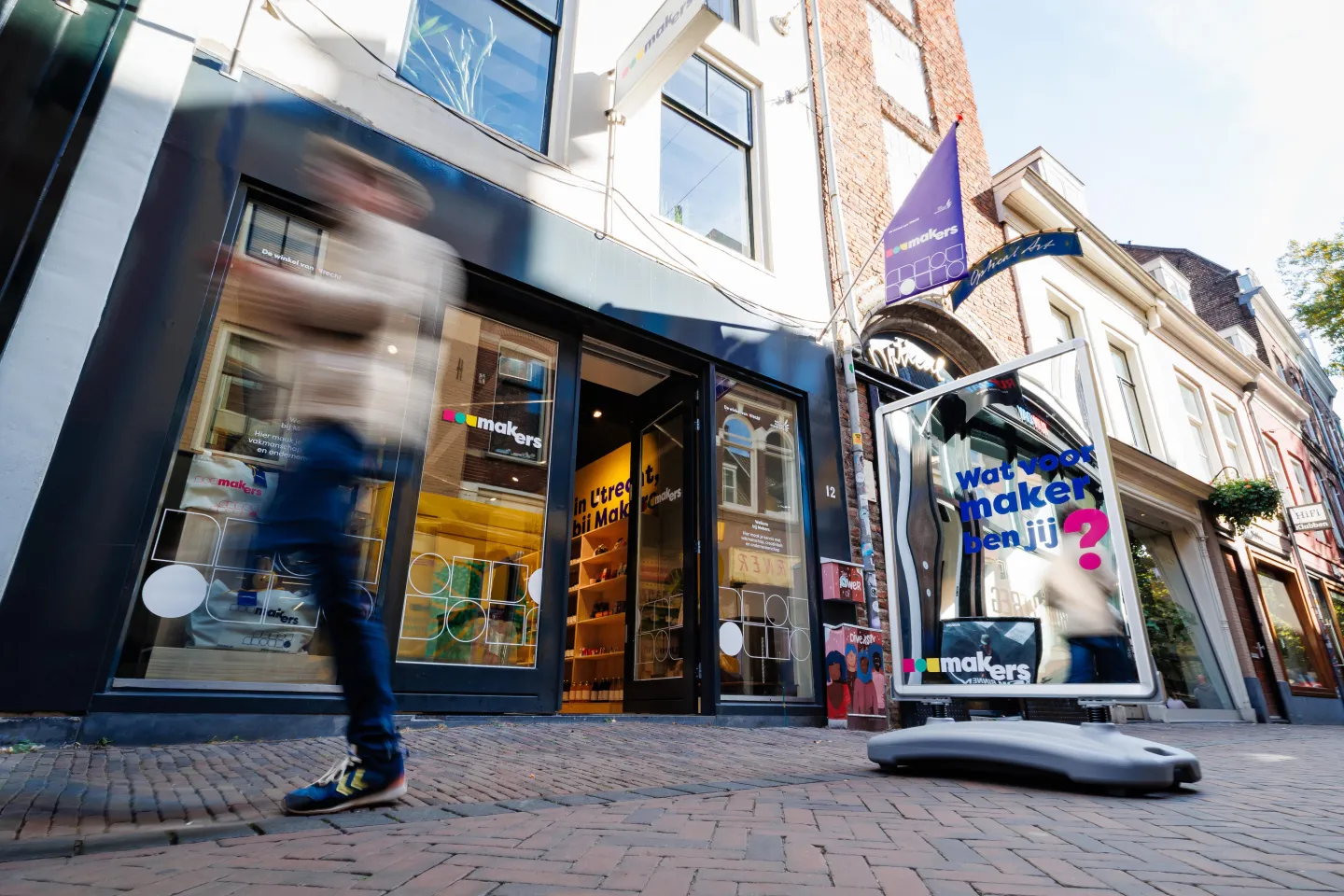 Buitenaanzicht van de Makerswinkel van ROC Midden Nederland in Utrecht, met open deur, uithangbord en stoepbord met de tekst “Wat voor maker ben jij?” terwijl een voorbijganger langsloopt.