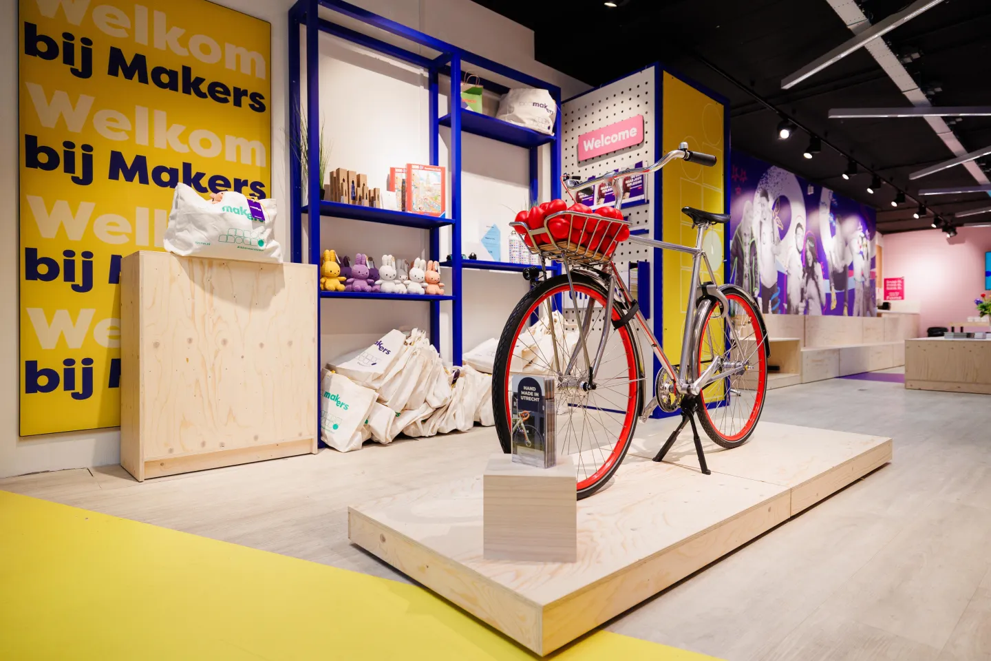 Fiets met rode wielen op een houten plateau in de Makerswinkel van ROC Midden Nederland, met op de achtergrond gele en paarse wanden, schappen met producten en tassen met het Makers-logo.