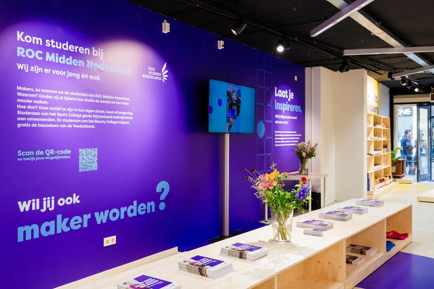 Interieuraanzicht van de Makerswinkel van ROC Midden Nederland met paarse wanden, informatie over opleidingen, een QR-code, stapels brochures en bloemen op houten tafels.