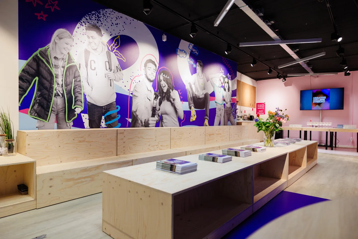 Interieur van de Makerswinkel van ROC Midden Nederland met houten zitblokken, stapels brochures, een bloemenvaas en een grote muur met zwart-witfoto’s van studenten op een paarse achtergrond.