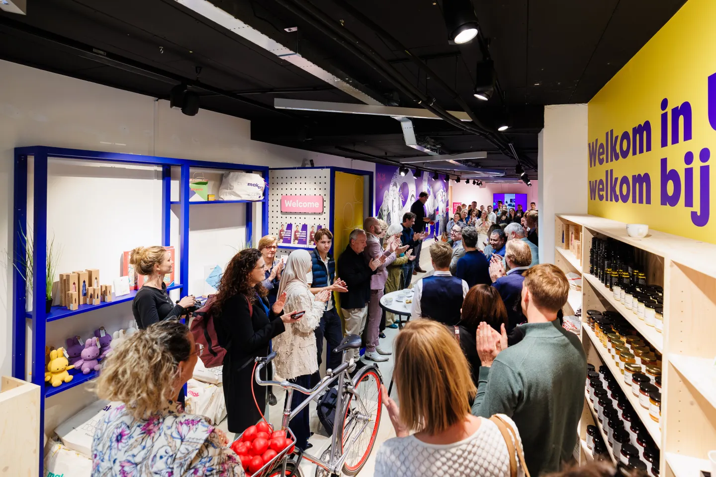 Drukke opening van de Makerswinkel van ROC Midden Nederland met bezoekers die applaudisseren, producten op houten en blauwe schappen en kleurrijke wanden met teksten als “Welkom bij de Makers”.