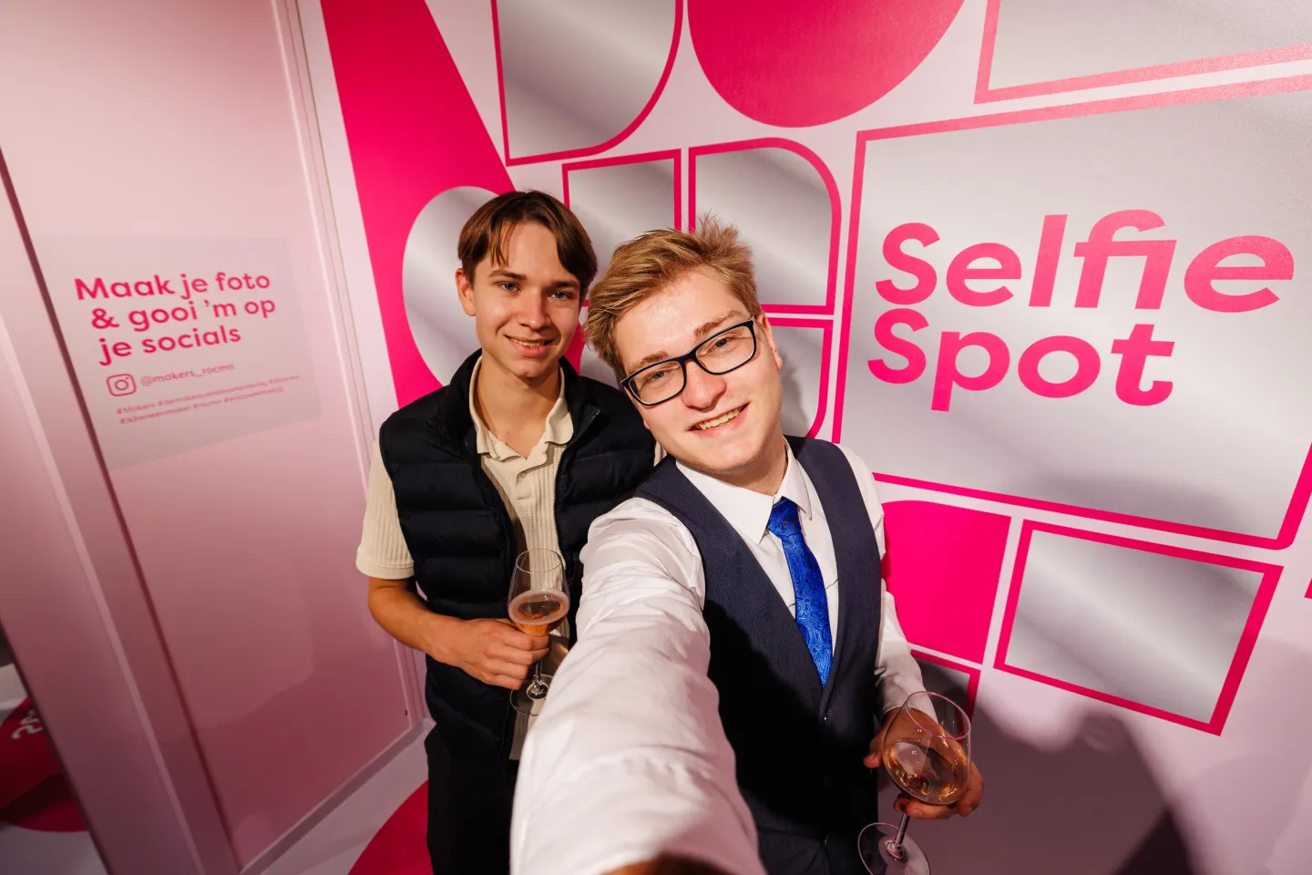 Twee bezoekers maken een selfie bij de roze Selfie Spot in de Makerswinkel van ROC Midden Nederland, met wijnglazen in de hand en een achtergrond met de tekst “Maak je foto & gooi ’m op je socials.