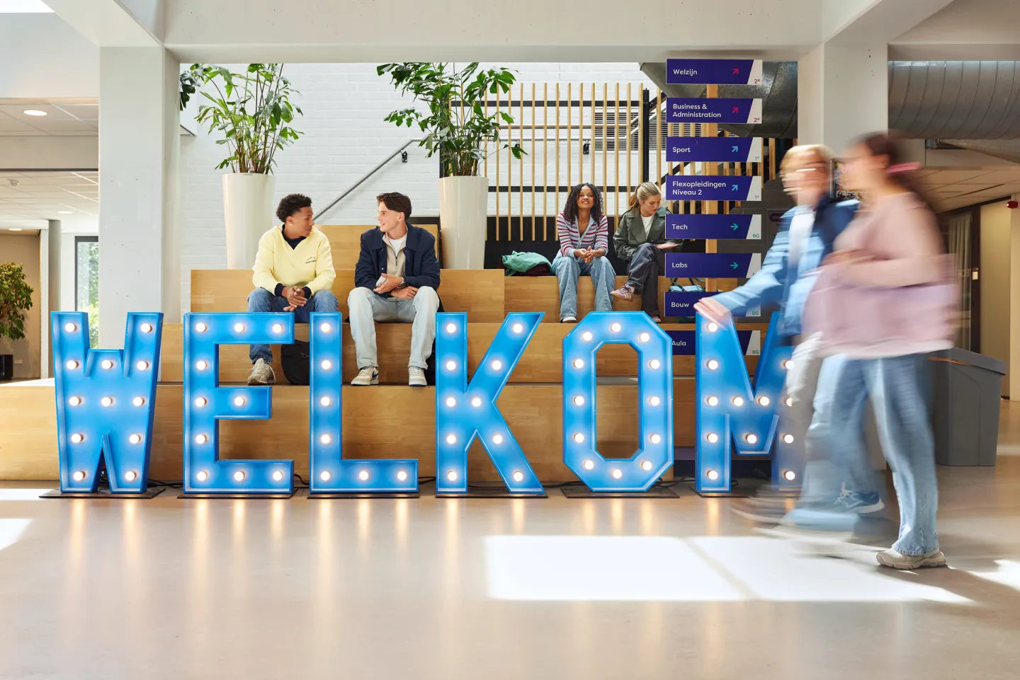 Studenten zitten bij een groot welkom-bord in de ingang van een schoolgebouw