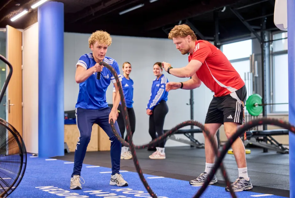Een fitnesstrainer coacht een sporter bij een oefening met zware lange touwen.