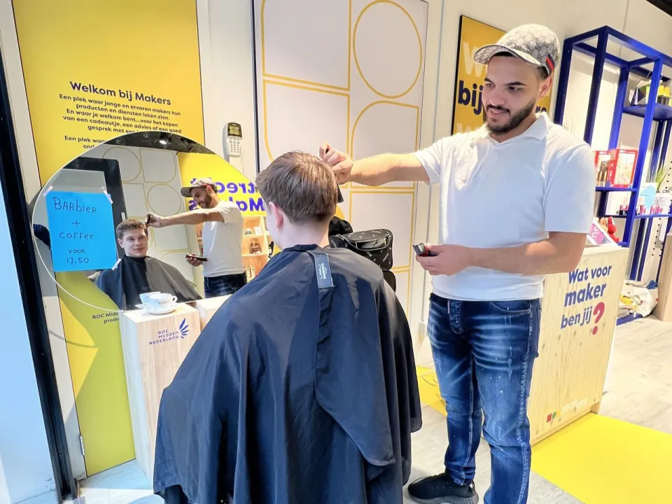 Kappersstudent die het haar knipt van een jongen in de Makers-omgeving, met spiegel en werkplek op de achtergrond.