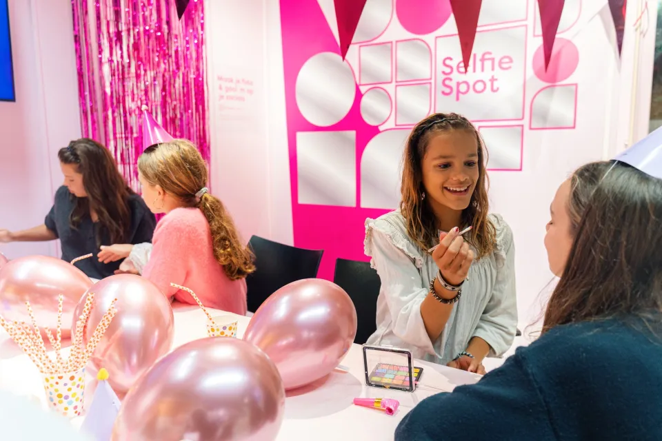 Kinderen brengen make-up aan bij elkaar tijdens een feestelijke kinderworkshop bij de Selfie Spot van ROC Midden Nederland, met roze ballonnen en feesthoedjes op tafel.