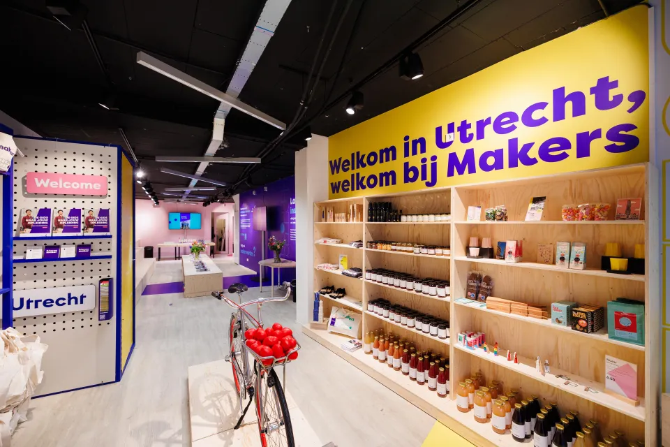 Overzicht van de Makerswinkel van ROC Midden Nederland in Utrecht met houten schappen vol producten, een rode fiets, paarse en gele wanden met teksten als “Welkom in Utrecht, welkom bij Makers”, en een moderne opstelling met informatiebalie en schermen.