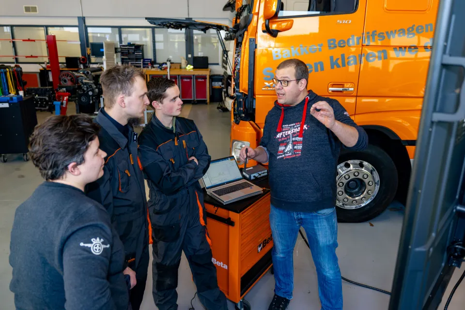 Docent legt een techniekopdracht uit aan drie studenten in een werkplaats bij een vrachtwagen.