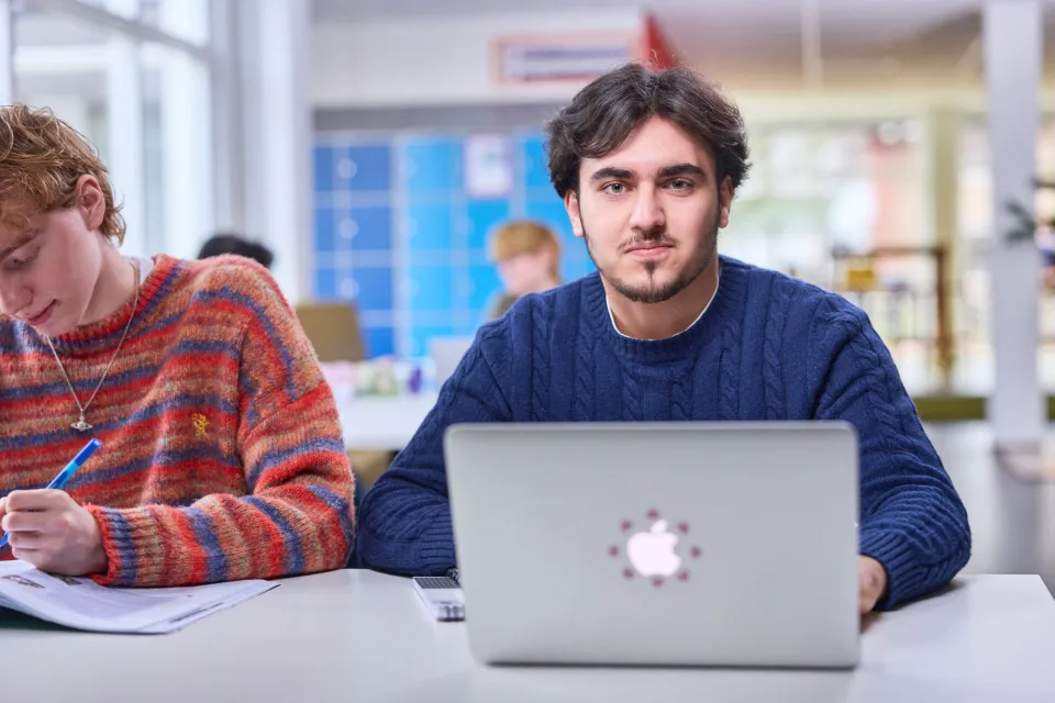 Student zit achter een laptop aan een tafel in een klas- of studieruimte, met een andere student die naast hem aantekeningen maakt.