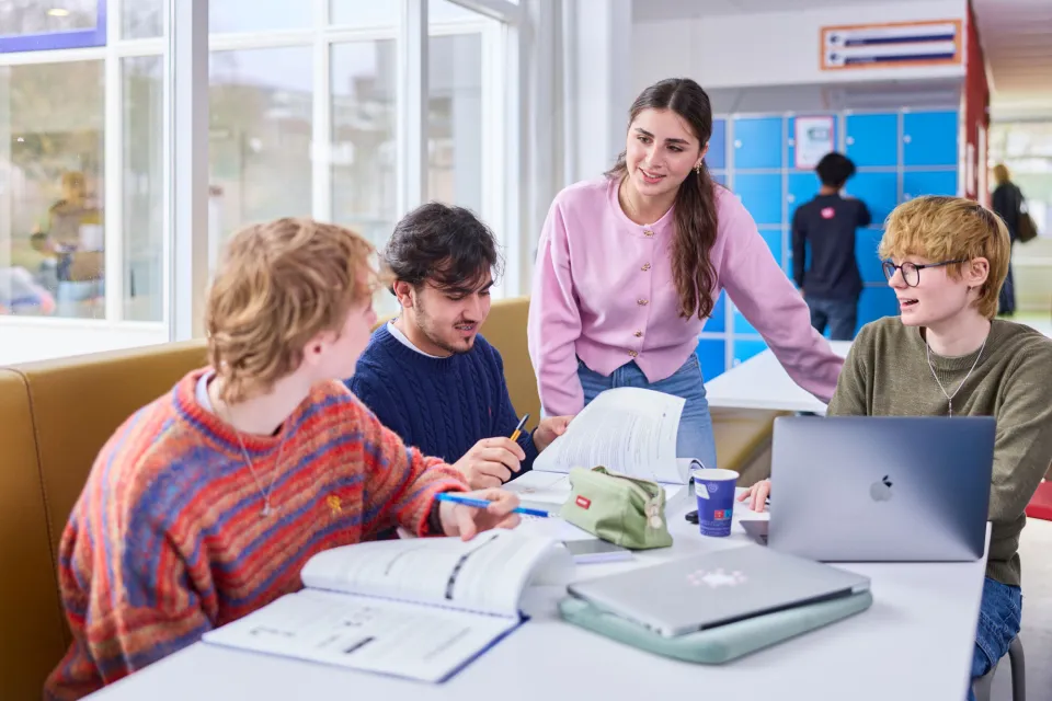 Docent staat bij een tafel en begeleidt vier studenten die samen aan het studeren zijn met boeken en laptops in een open leeromgeving.