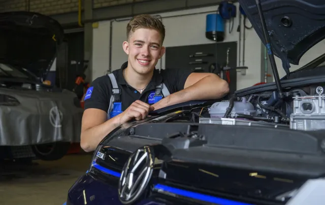 Student glimlacht terwijl hij voor een Volkswagen auto in de werkplaats zit.