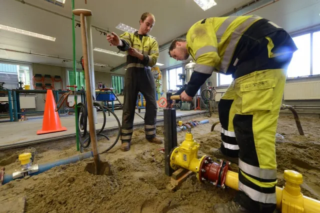 Twee monteurs gas-, water- en warmtedistributie werken aan een leiding op een bouwplaats.