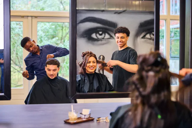 Studenten kappen klanten in een kapsalon tijdens praktijkles, met spiegels en vrolijke sfeer.