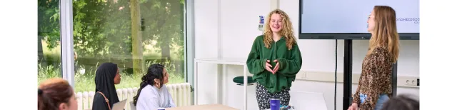 Studenten presenteren in de klas; een student vertelt enthousiast voor de groep terwijl klasgenoten luisteren en aantekeningen maken met laptops open.