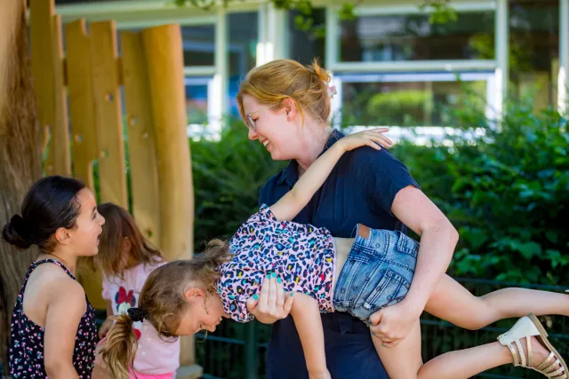 Een vrouw houdt een klein meisje in de lucht. Een vrolijke en speelse interactie tussen een begeleider en kinderen op een speelplaats.