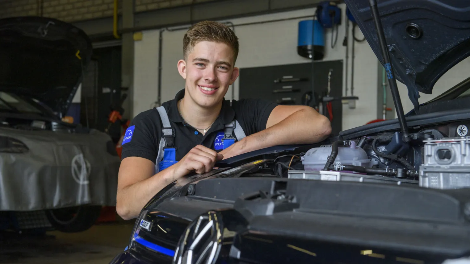 Student glimlacht terwijl hij voor een Volkswagen auto in de werkplaats zit.