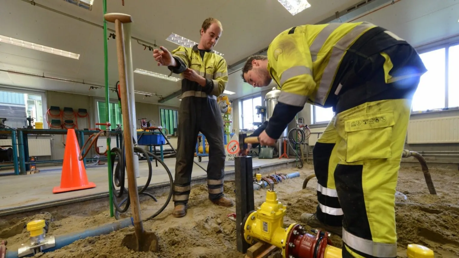 Twee monteurs gas-, water- en warmtedistributie werken aan een leiding op een bouwplaats.