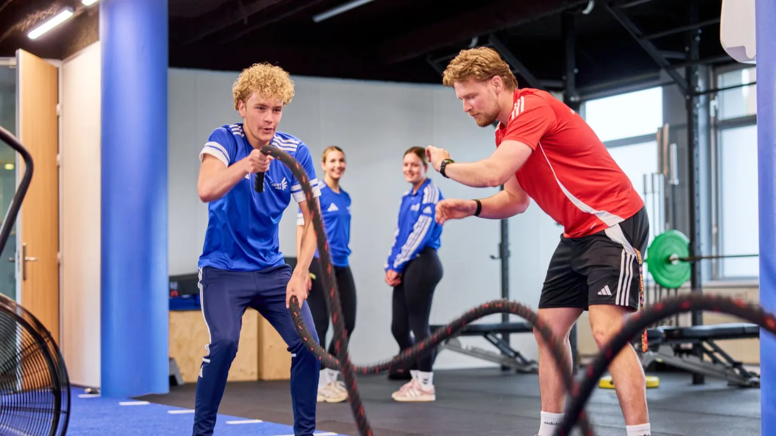 Een fitnesstrainer coacht een sporter bij een oefening met zware lange touwen.
