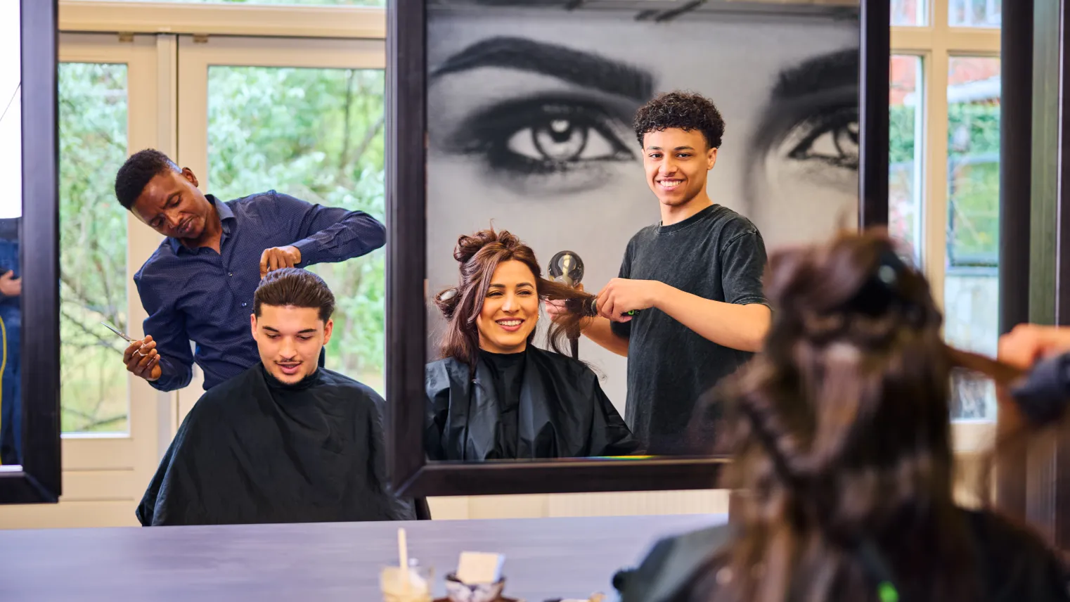 Studenten kappen klanten in een kapsalon tijdens praktijkles, met spiegels en vrolijke sfeer.