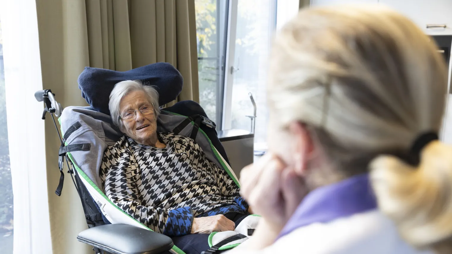 Een oudere vrouw in een rolstoel kijkt naar de zorgmedewerker tegenover haar.