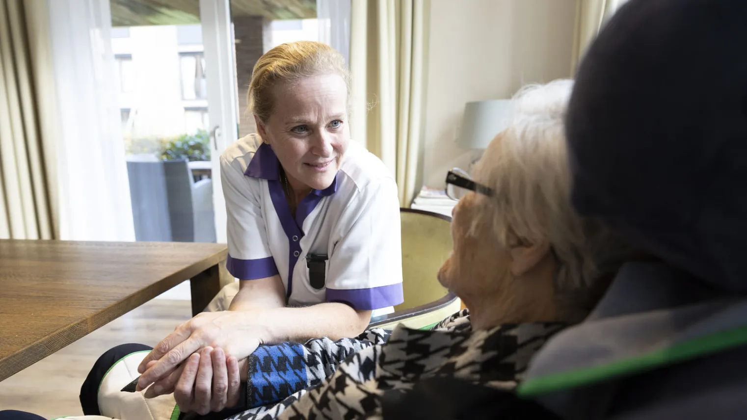 Een zorgmedewerkers houdt de hand vast van een oudere vrouw in een rolstoel.