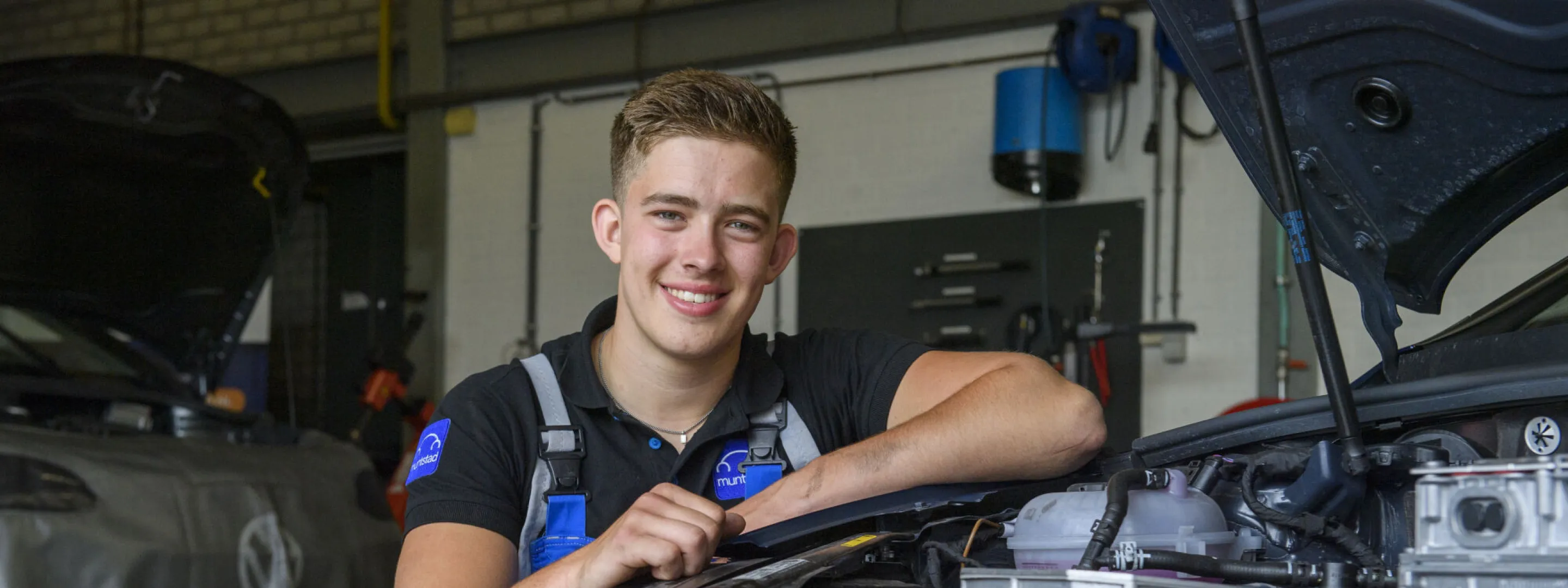 Student glimlacht terwijl hij voor een Volkswagen auto in de werkplaats zit.