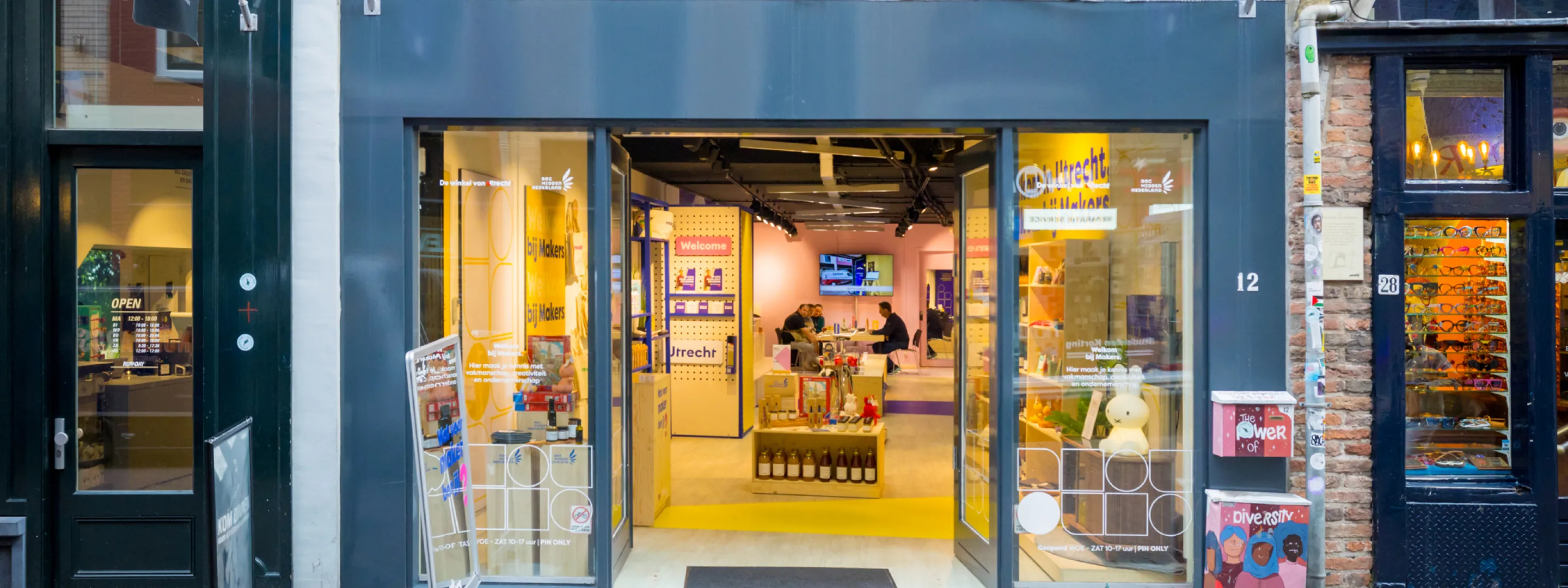 Ingang van de winkel Makers aan een gezellige winkelstraat in Utrecht. De glazen deuren staan open en binnen zijn houten displays met lokale producten te zien. Aan de gevel hangen blauwe vlaggen met het Makers-logo, en voor de deur staan reclameborden op de stoep.