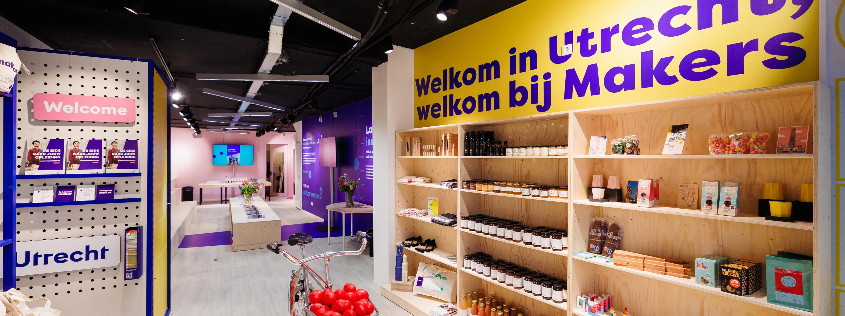 Overzicht van de Makerswinkel van ROC Midden Nederland in Utrecht met houten schappen vol producten, een rode fiets, paarse en gele wanden met teksten als “Welkom in Utrecht, welkom bij Makers”, en een moderne opstelling met informatiebalie en schermen.