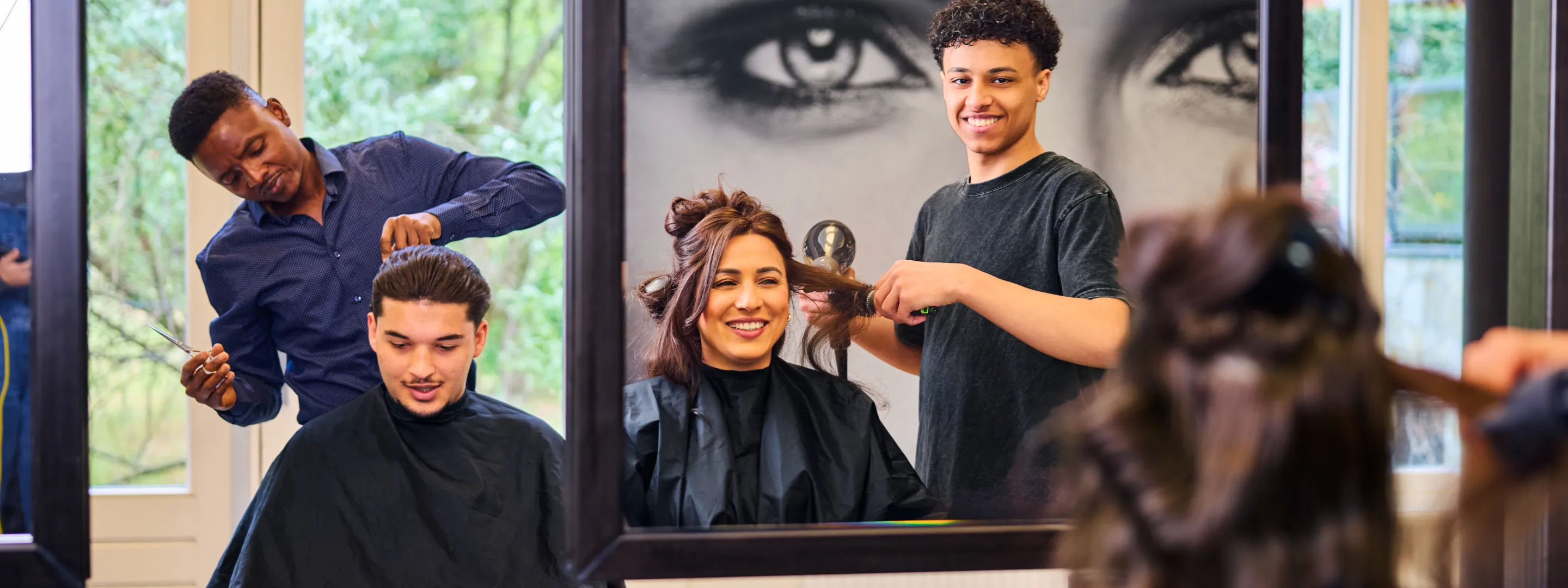 Studenten kappen klanten in een kapsalon tijdens praktijkles, met spiegels en vrolijke sfeer.