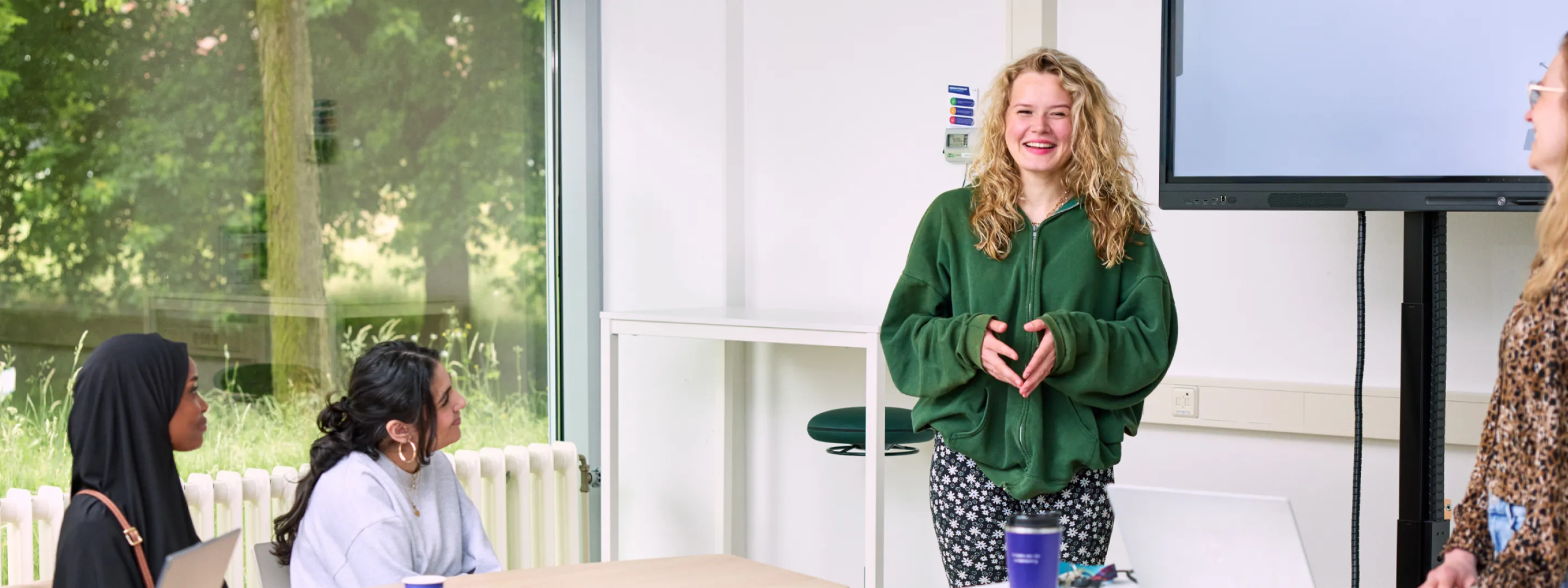 Studenten presenteren in de klas; een student vertelt enthousiast voor de groep terwijl klasgenoten luisteren en aantekeningen maken met laptops open.
